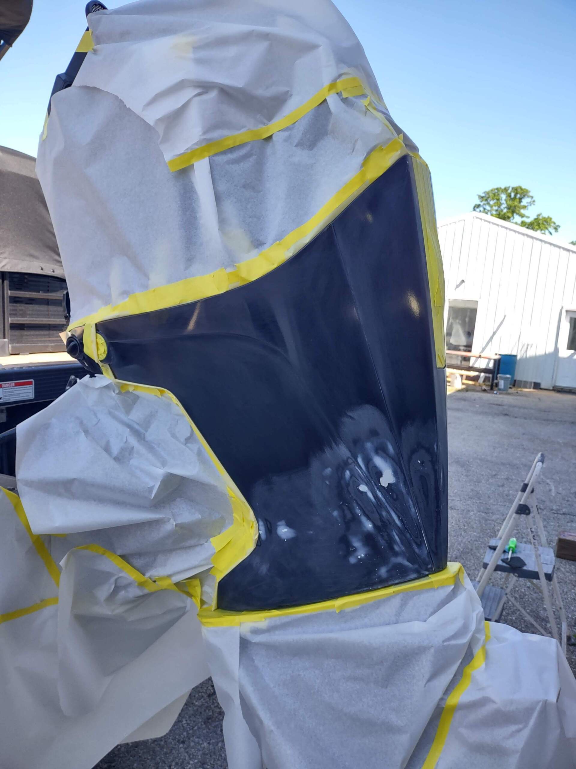 A masked person in a white hooded suit with yellow tape stands outdoors near industrial buildings.