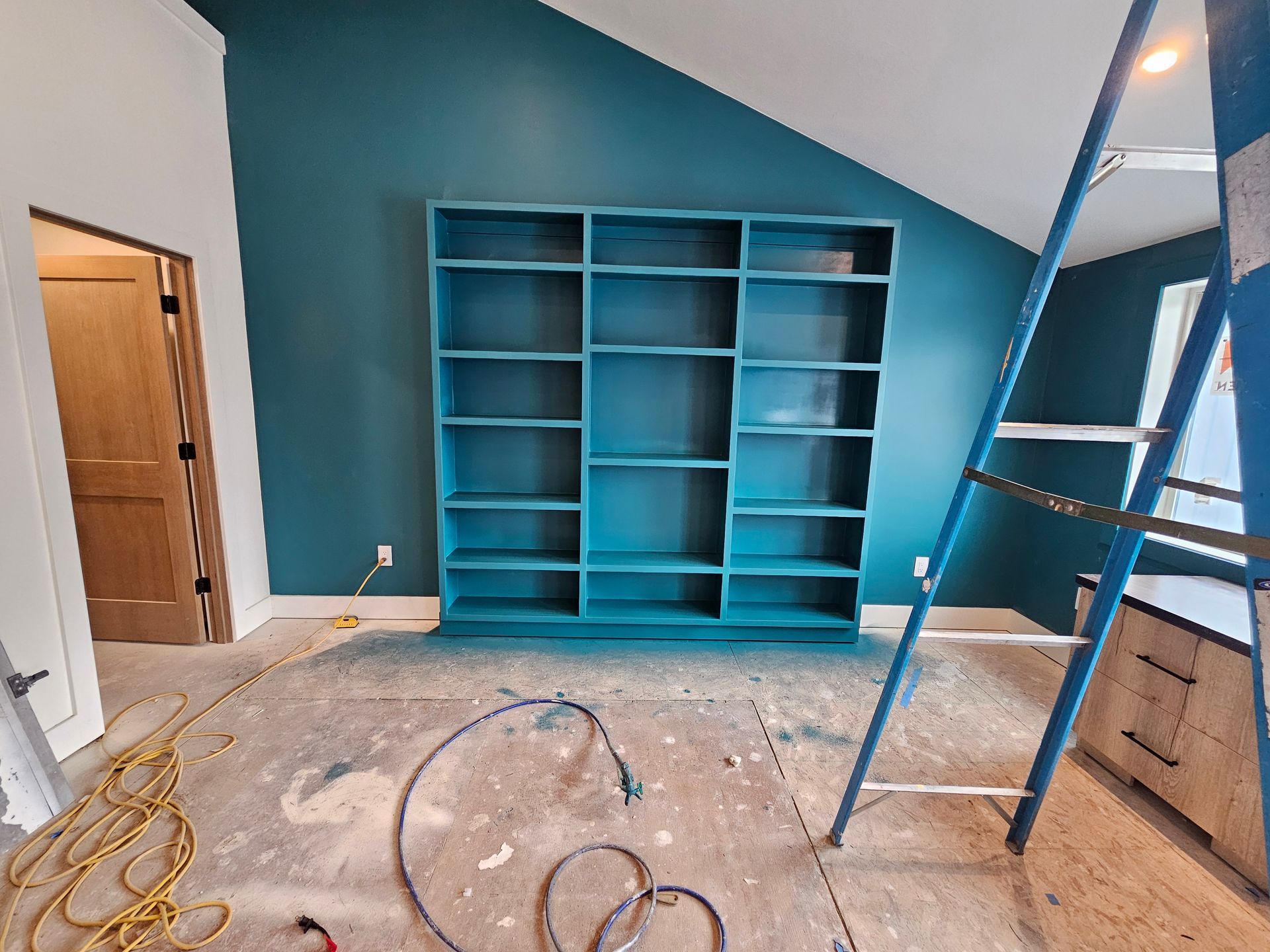 Teal bookshelf against a teal wall; ladder, open door, and exposed floorboards present.