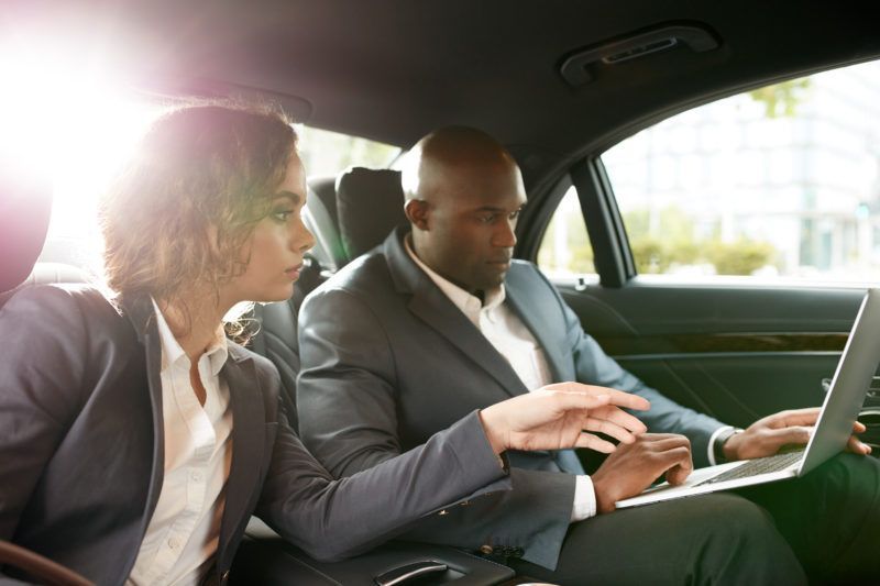 A man and a woman are sitting in the back seat of a car looking at a laptop.
