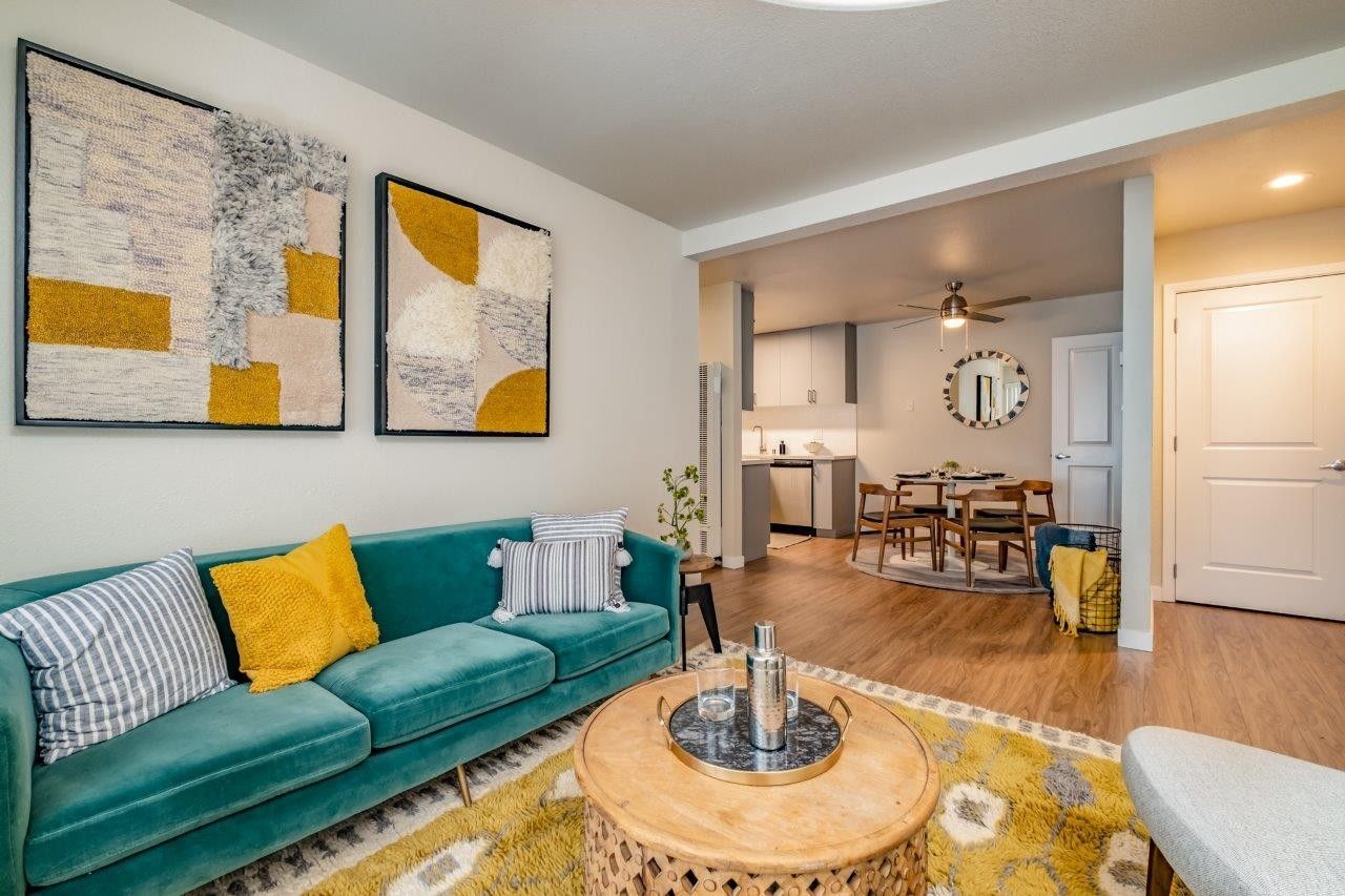 Modern living room with teal sofa, yellow accents, wall art, and open view to dining area and kitchen.