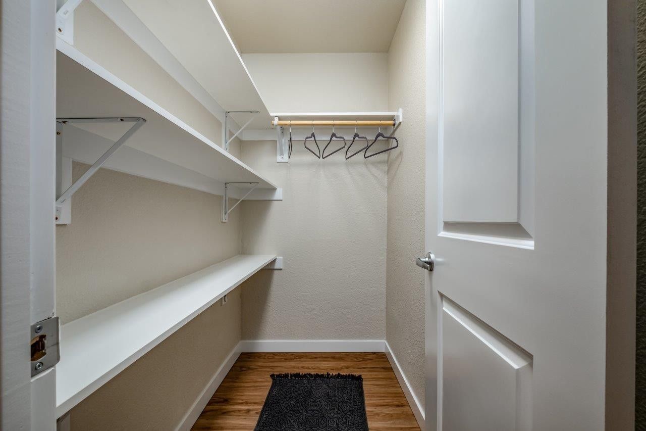 Empty walk-in closet with white shelves, hanging rods, and a black rug on wood flooring.