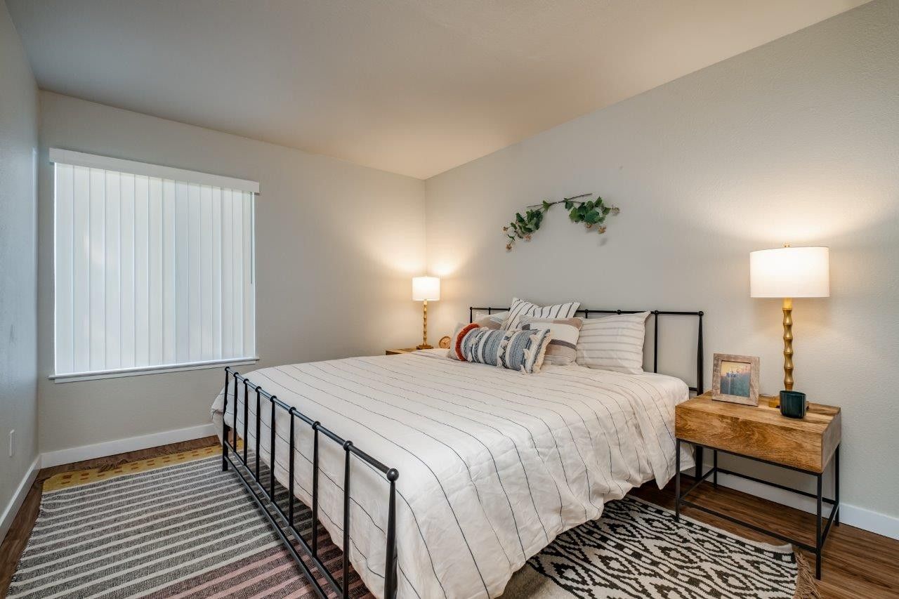 Bright bedroom with a black metal bed, nightstands, lamps, white walls, and a window with blinds.