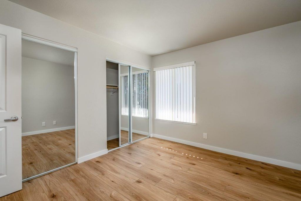 Empty bedroom with light gray walls, hardwood floors, a window, and mirrored closet doors
