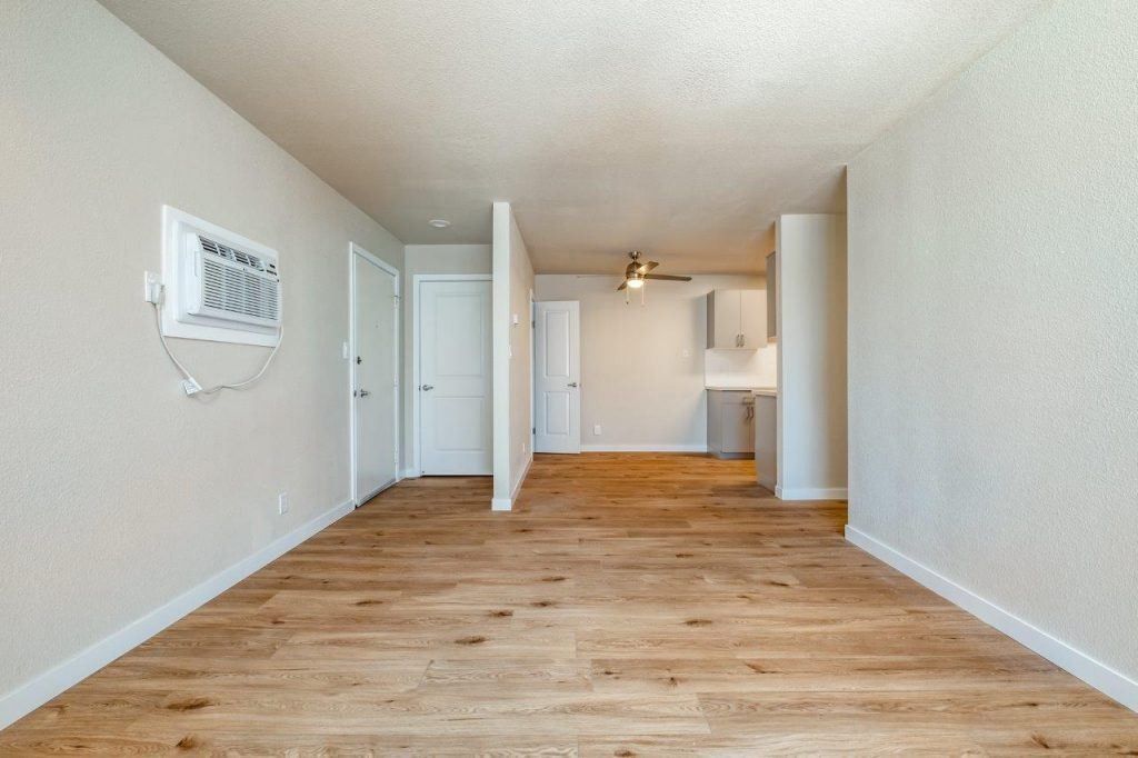 Empty beige apartment room with wood floors, wall AC unit, and open kitchen in the back
