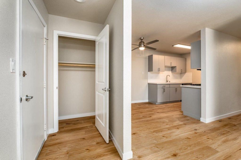 Empty hallway with open closet, white walls, and a view into a kitchen with wood floors.