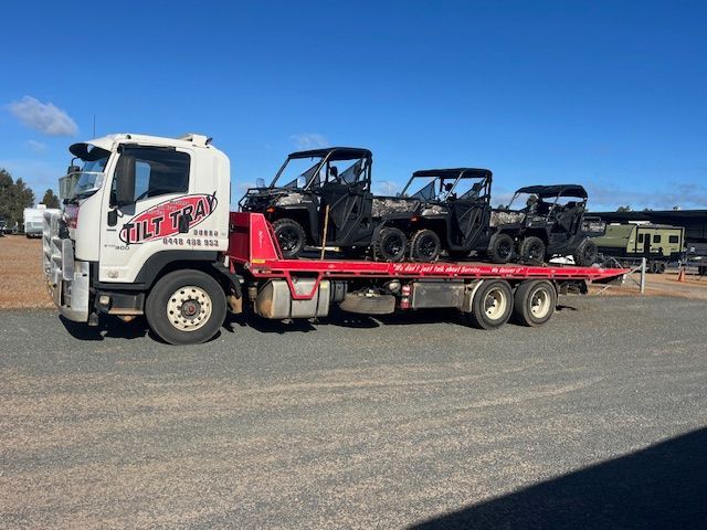 White tilt tray truck carrying four black side-by-side utility vehicles on a flatbed.