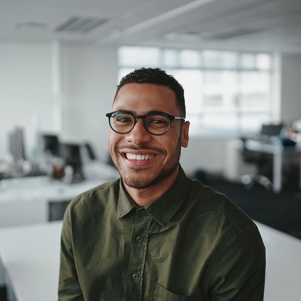 A man wearing glasses and a green shirt is smiling in an office.