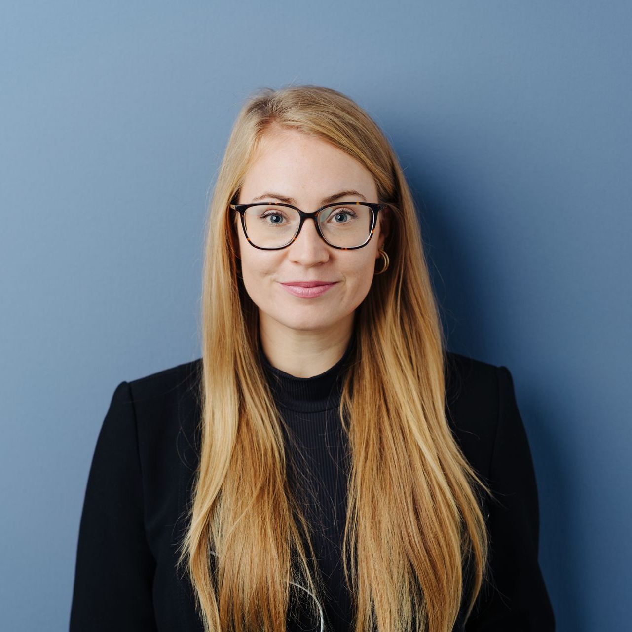 A woman wearing glasses and a black jacket is standing in front of a blue wall.