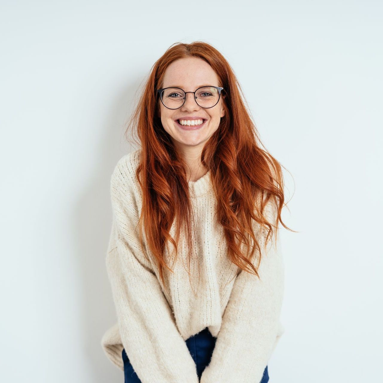 A woman with red hair and glasses is wearing a white sweater and smiling.