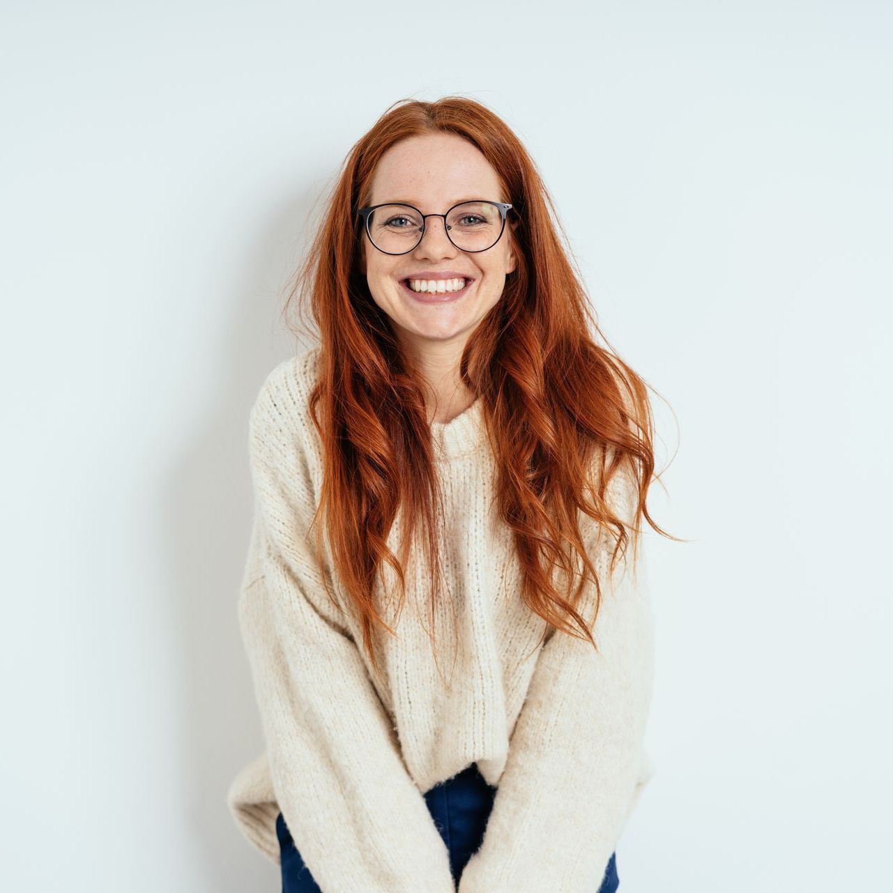 A woman with red hair and glasses is wearing a white sweater and smiling.