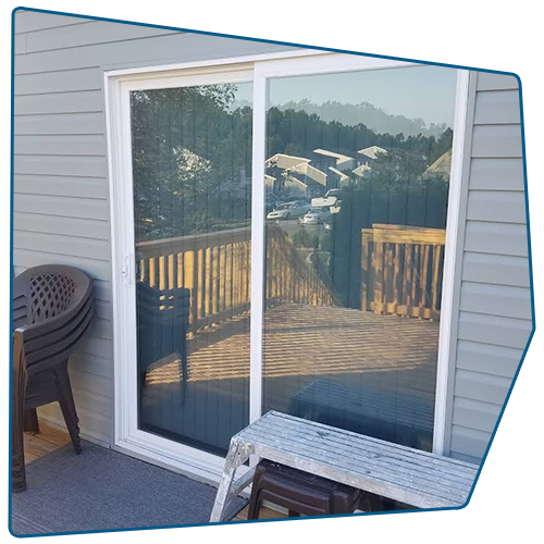 Sliding glass door on a deck, reflecting trees and buildings; chairs and a step stool are nearby.