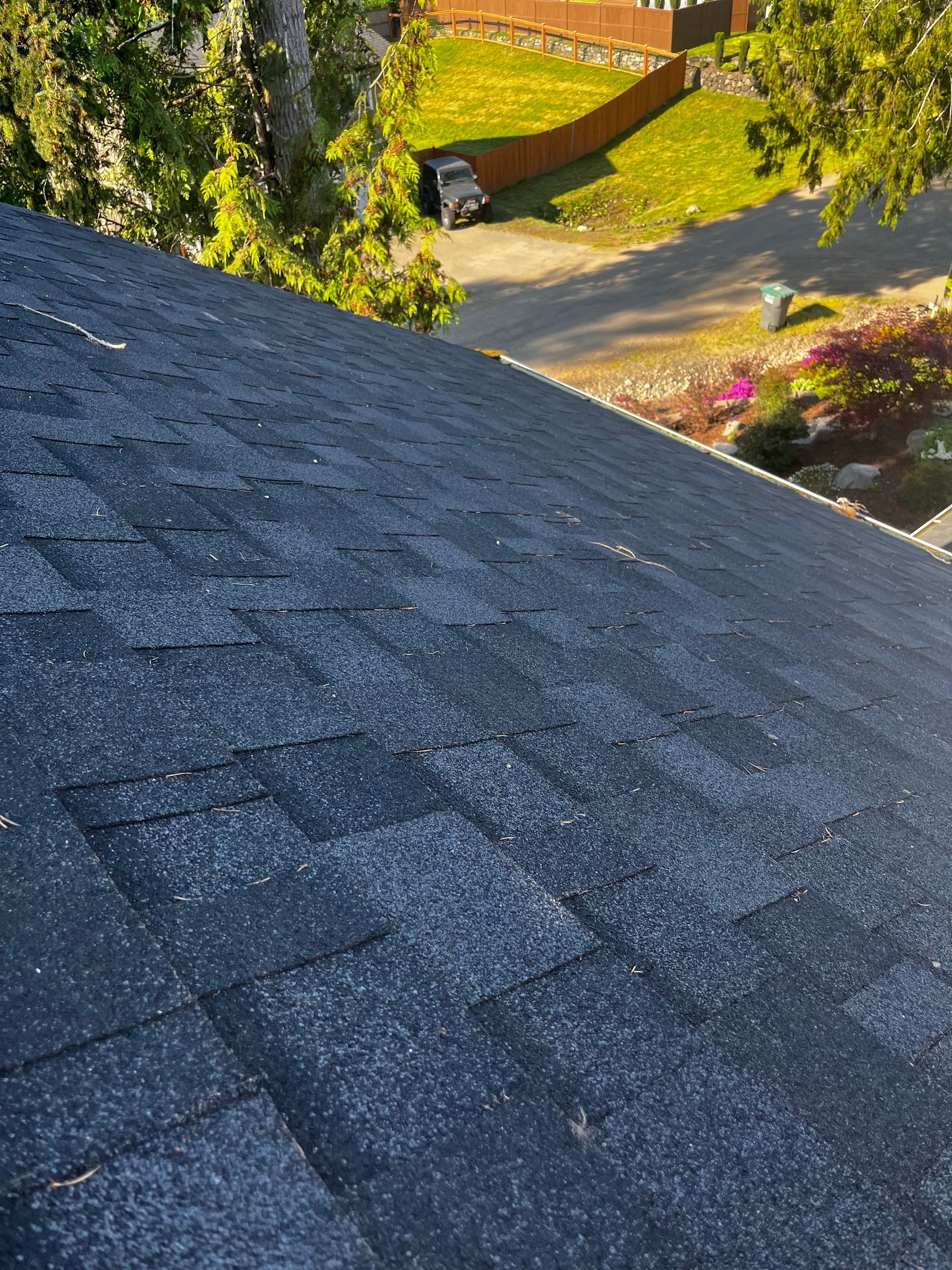Close-up of a dark gray shingled roof with a sunny yard and garden in the background