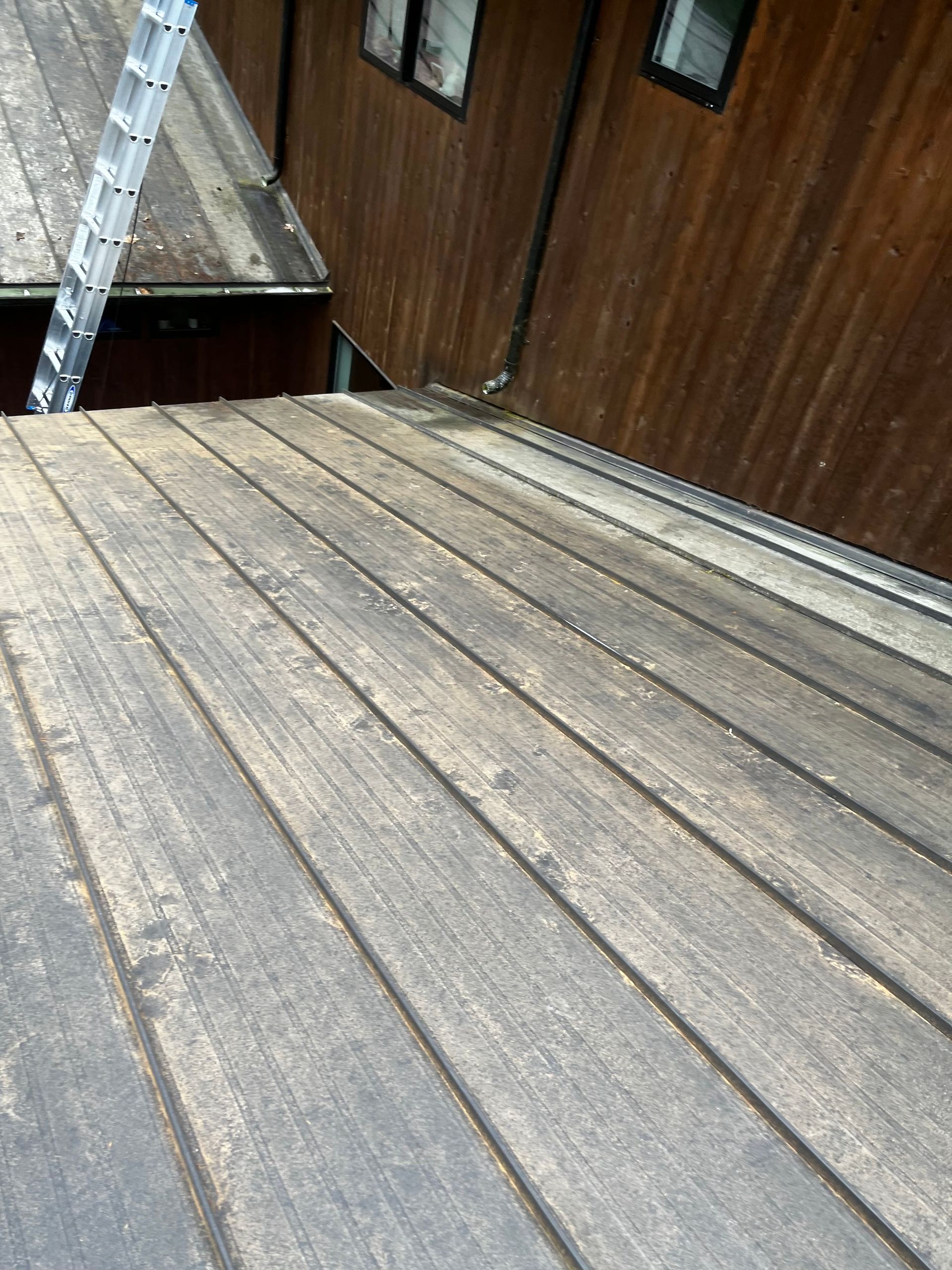 Weathered wooden deck beside a brown siding wall, with a ladder leaning on the left.