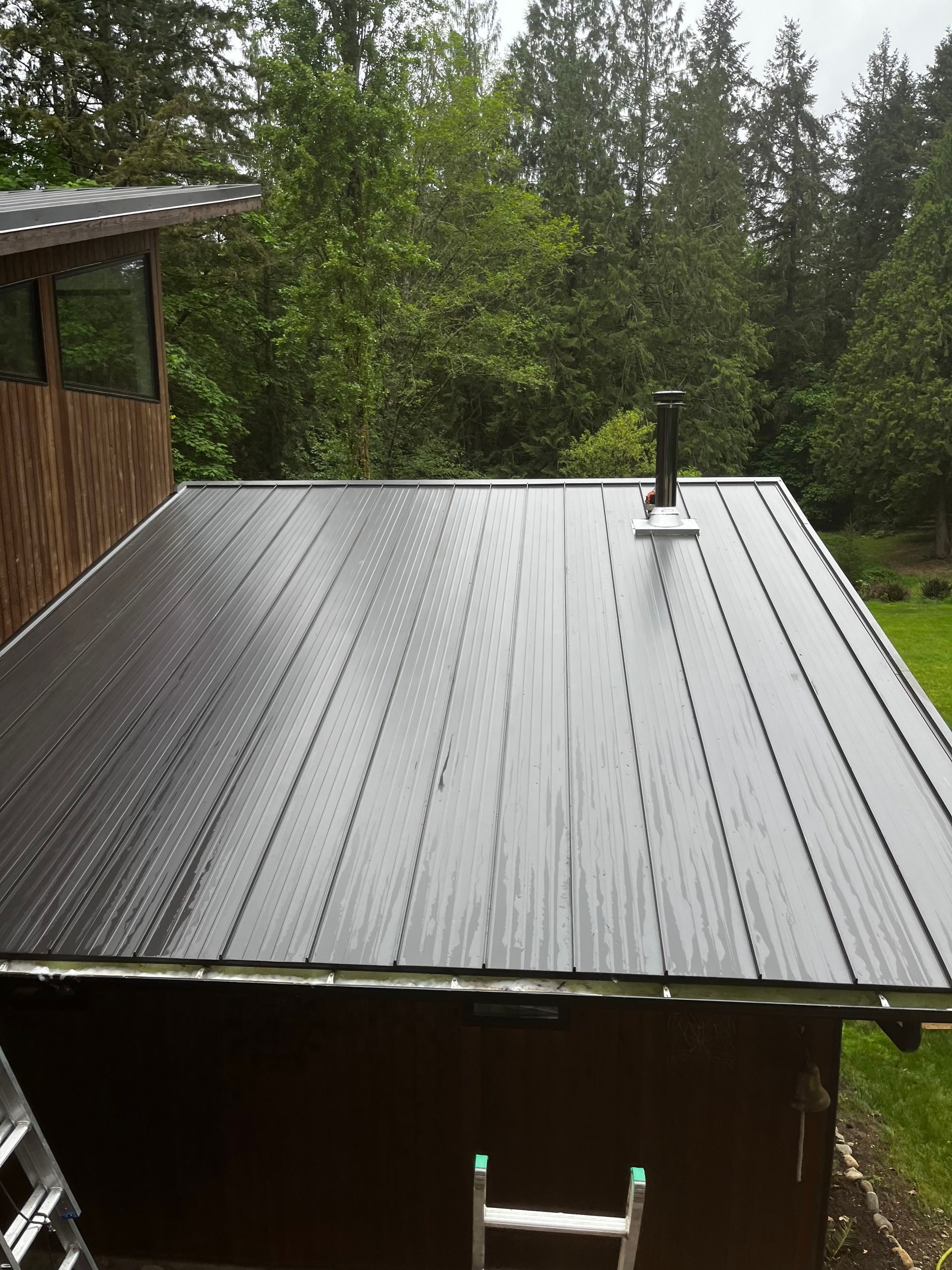 Rainy rooftop with dark metal panels, a small vent, and forest trees in the background