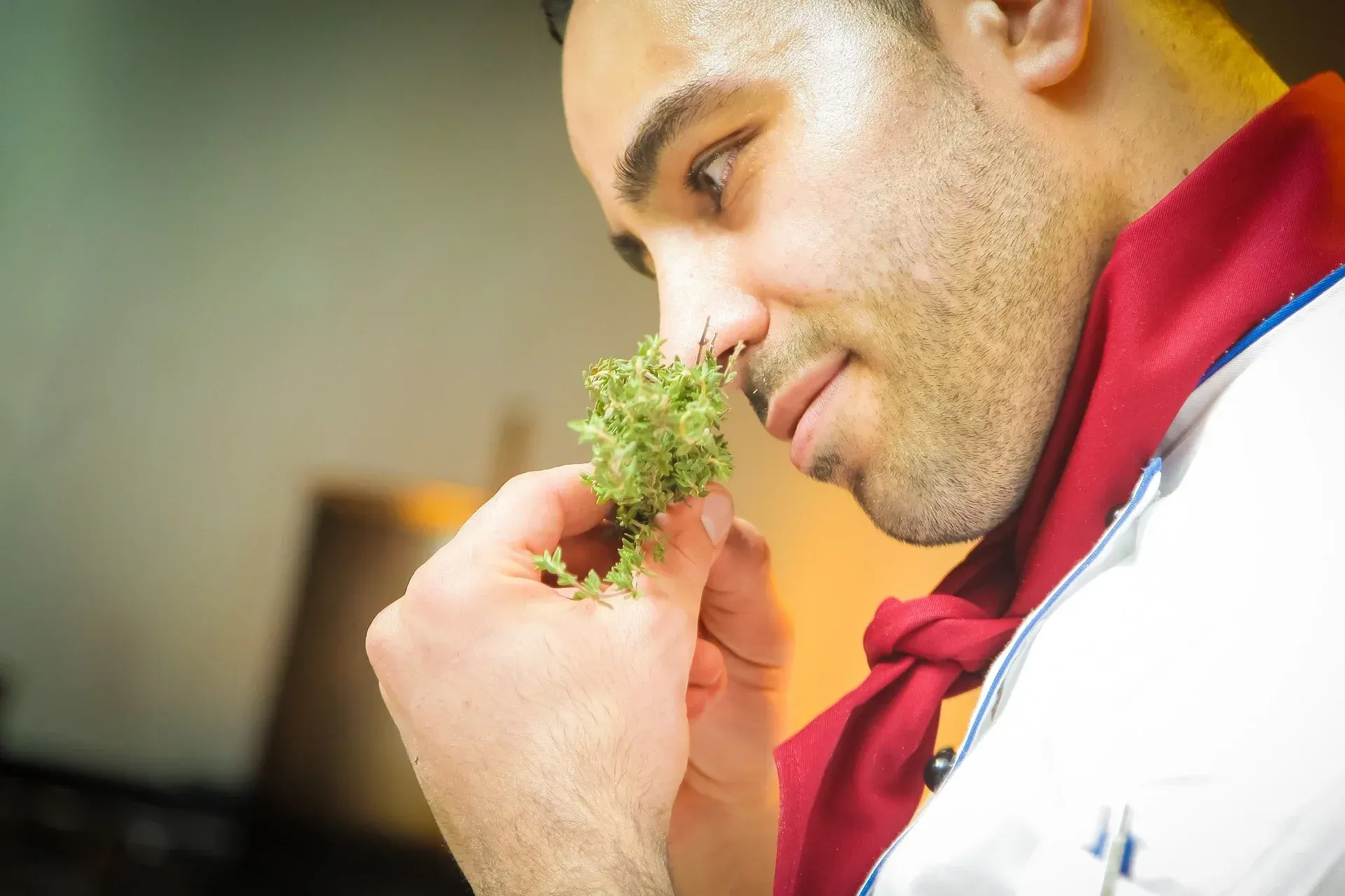A man in a chef 's uniform is smelling a green plant.