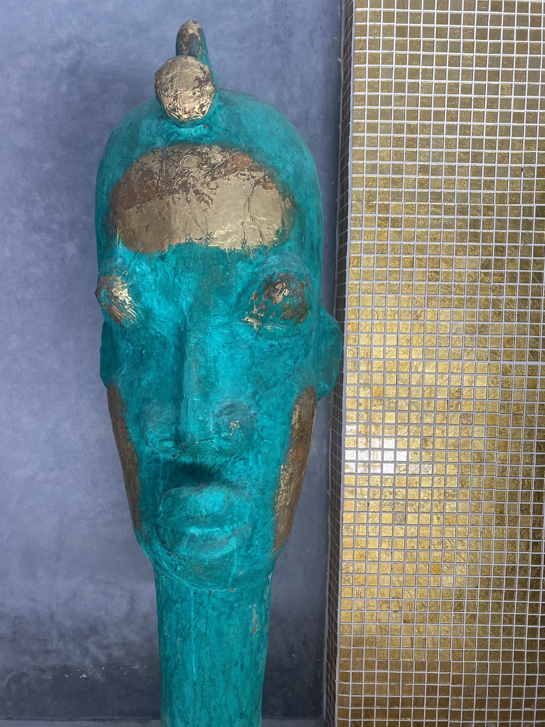 A blue and gold statue of a man 's head is sitting next to a gold mosaic wall.
