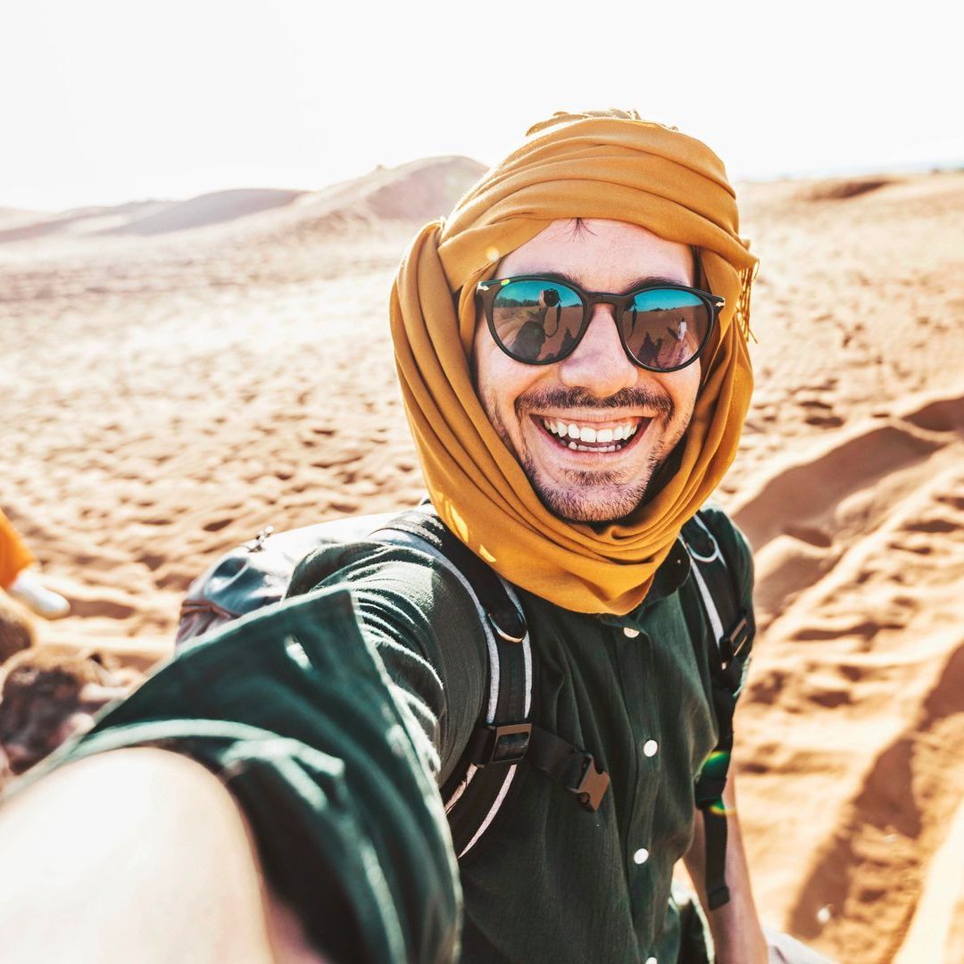 A man wearing sunglasses and a turban is taking a selfie in the desert.