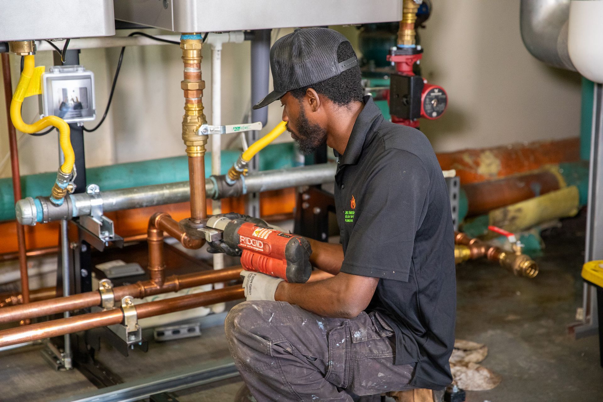 A man in work clothes kneels, soldering copper pipes in an industrial setting with various pipes and equipment.