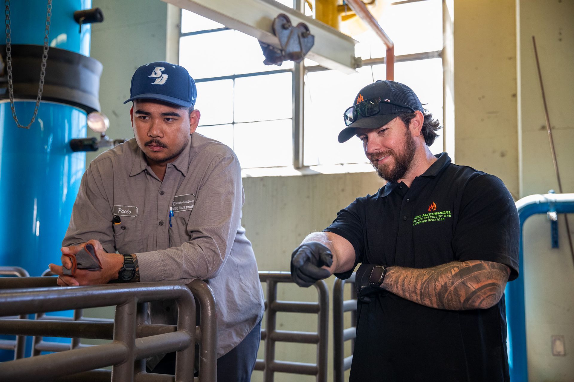 Two men in a factory setting; one wearing a work uniform and hat, the other in a black Jax Mechanical shirt, pointing.