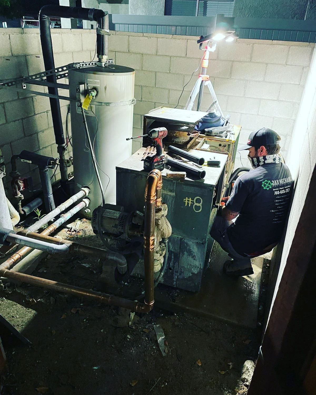 A technician works on a boiler in an outdoor utility space, lit by a work light. A water heater is next to the boiler.
