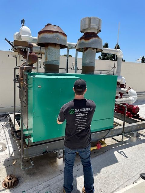 A man in jeans and a black Jax Mechanical t-shirt inspects a green rooftop generator unit.