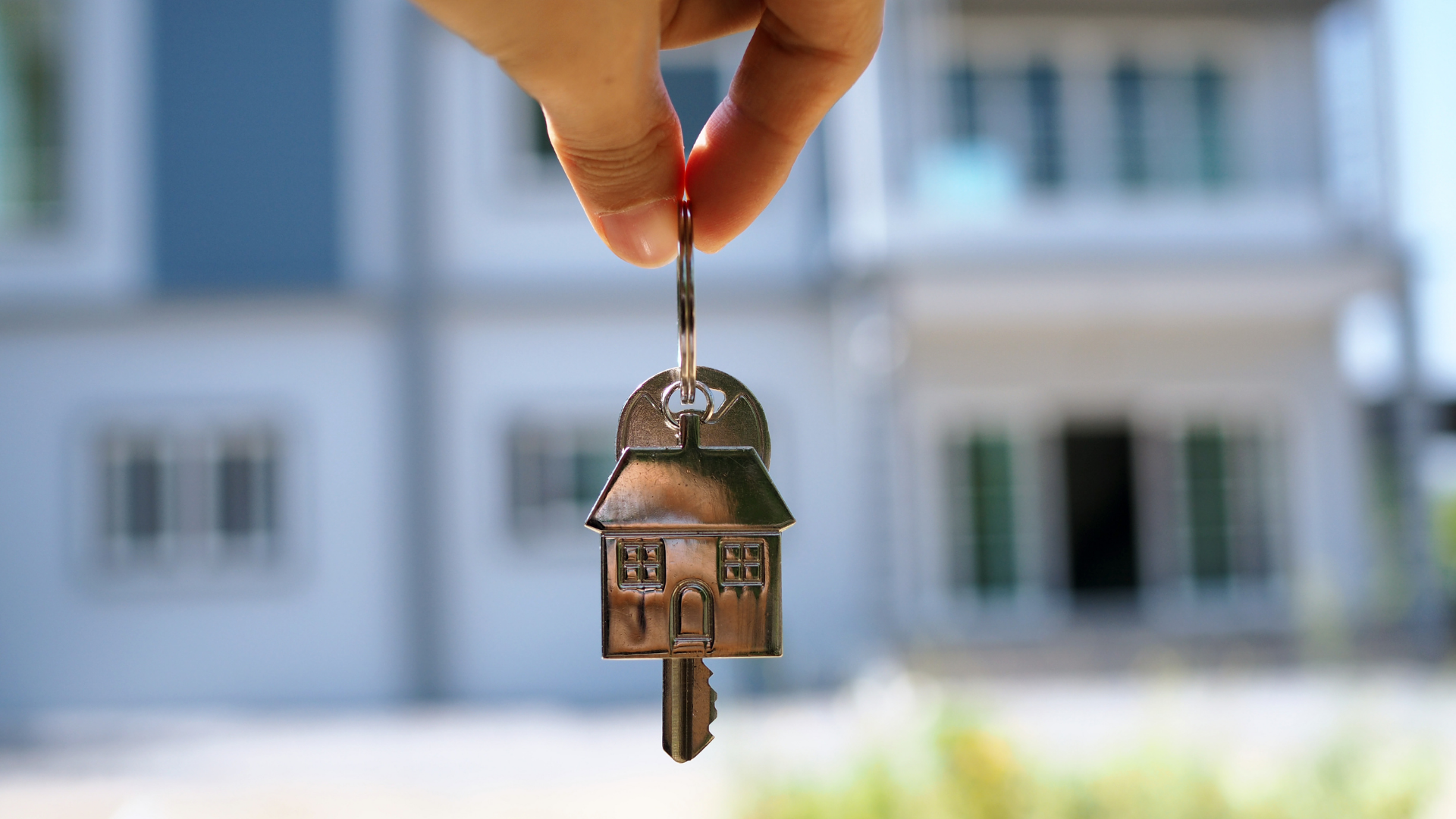 Hand holding house key in front of a house.