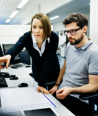 Woman pointing at a computer screen, discussing it with a man sitting beside her in an office setting.
