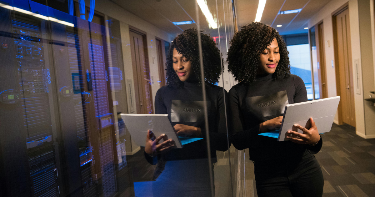 Woman with curly hair, holding a laptop, leans against a glass wall in an office setting.