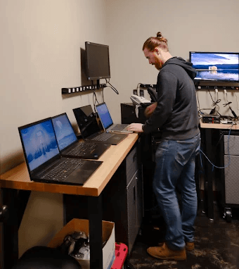 Person standing at a workstation with multiple monitors in a small office or studio.