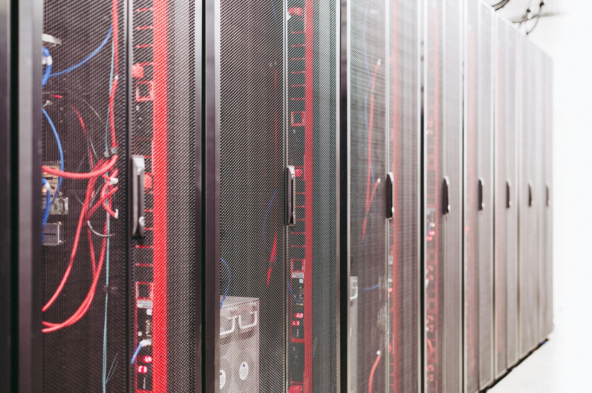 A row of black server racks in a data center, featuring visible red and blue cables through the mesh doors.