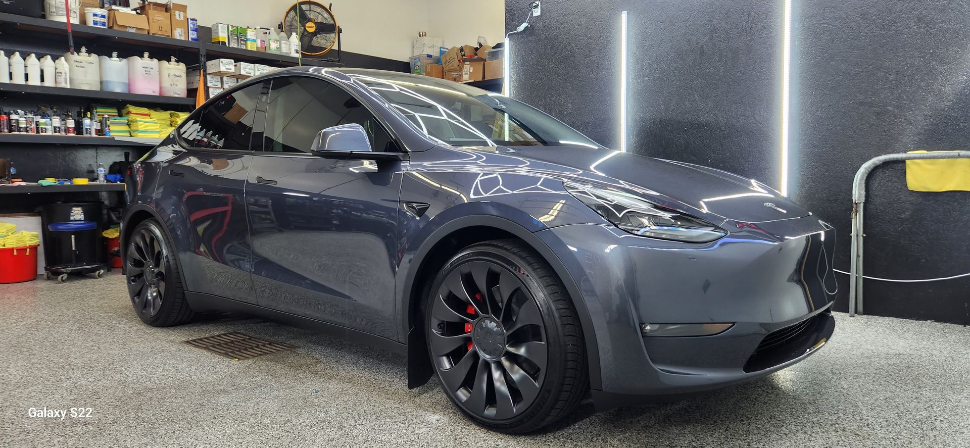 Dark gray Tesla parked in a garage, side view with black wheels and shop shelves in the background