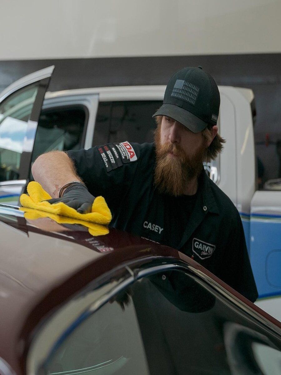 A man with a beard in a cap polishes a burgundy car roof with a yellow cloth. He's wearing black gloves and a work shirt.