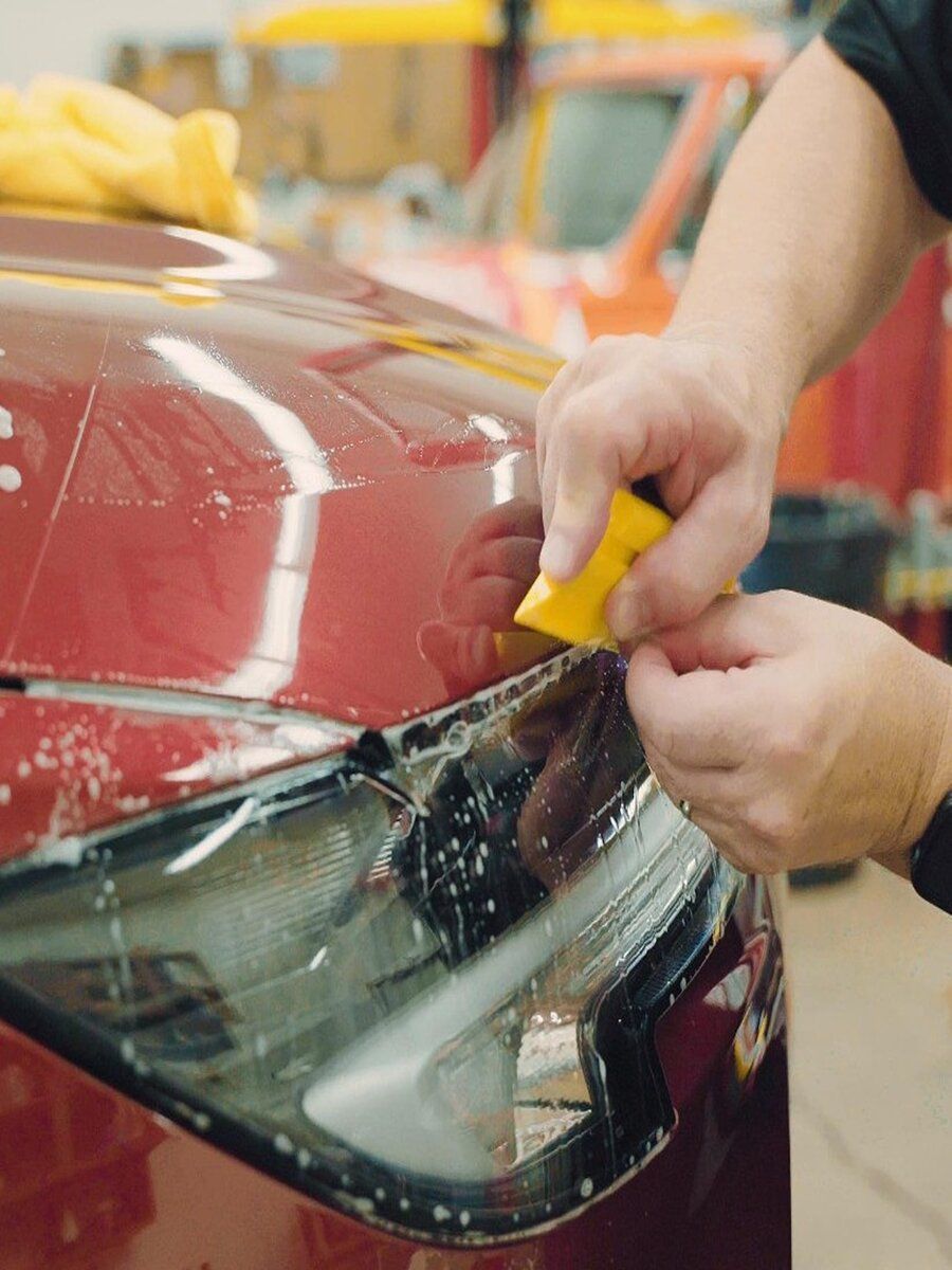 Person using a yellow squeegee to apply clear film to a car headlight. Red car in a workshop setting.