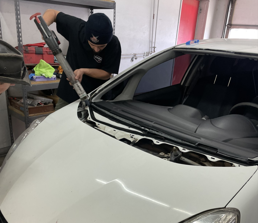Technician prying windshield from a white car in a workshop with tools and shelves nearby