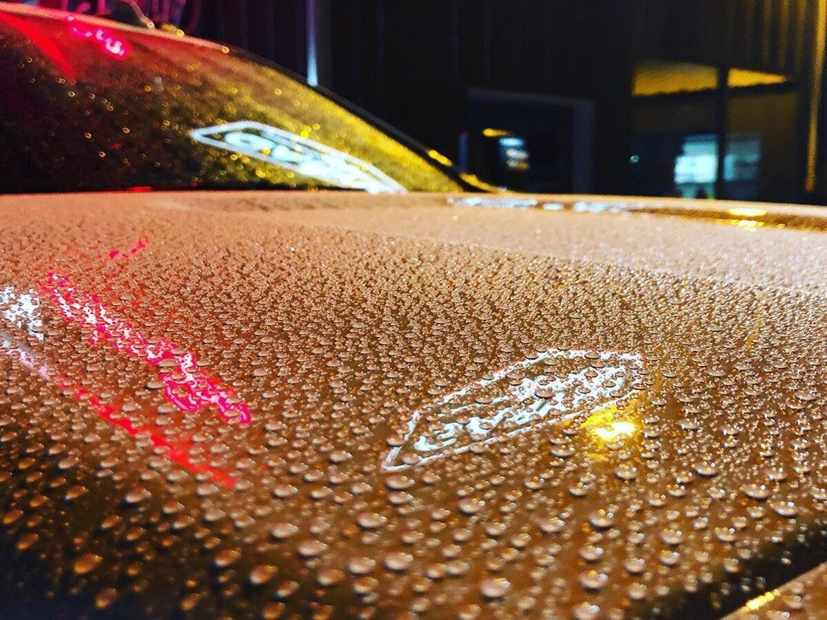 Close-up of a car hood covered in raindrops at night, with a logo partially visible. Red and white lights are reflected.