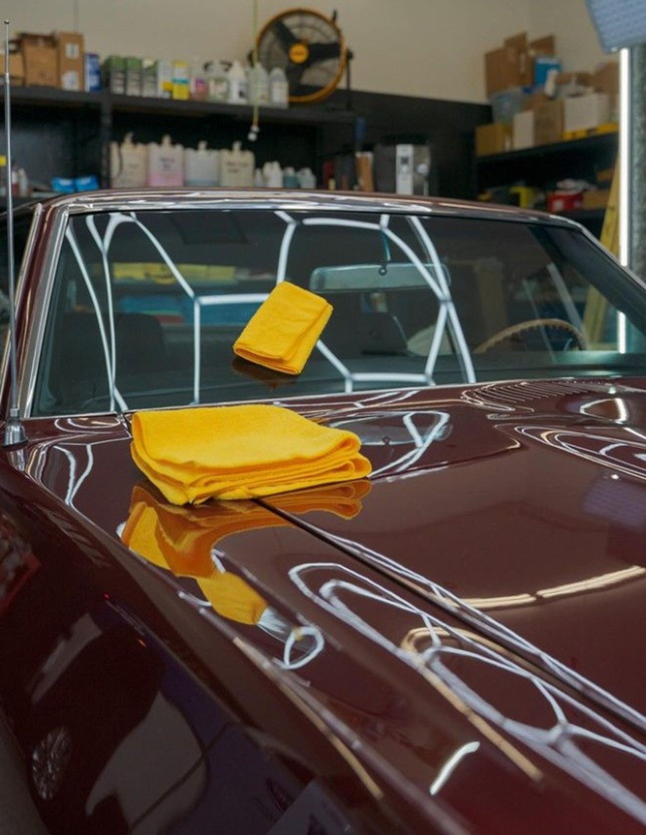 A shiny maroon classic car with yellow microfiber cloths on the hood, detailing shop in background.