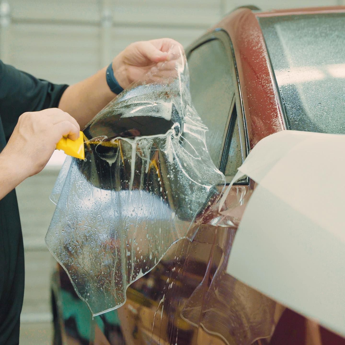 Person applying protective film to a red car side mirror with a yellow squeegee. Garage setting.