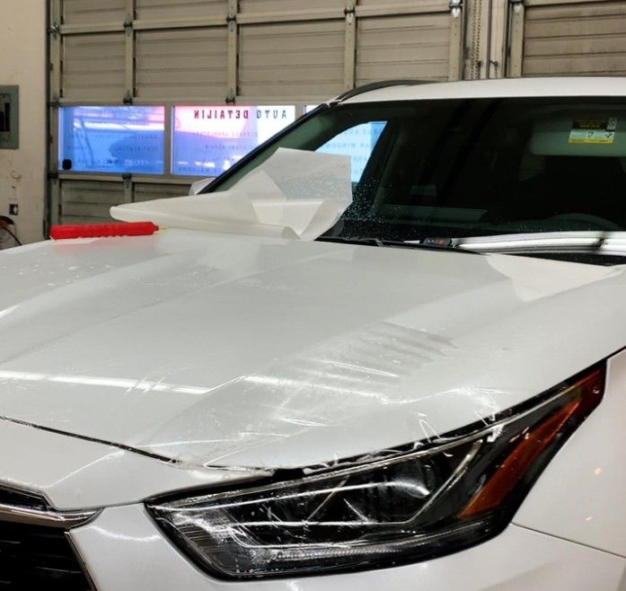 White SUV with clear film being applied to its hood in a garage.