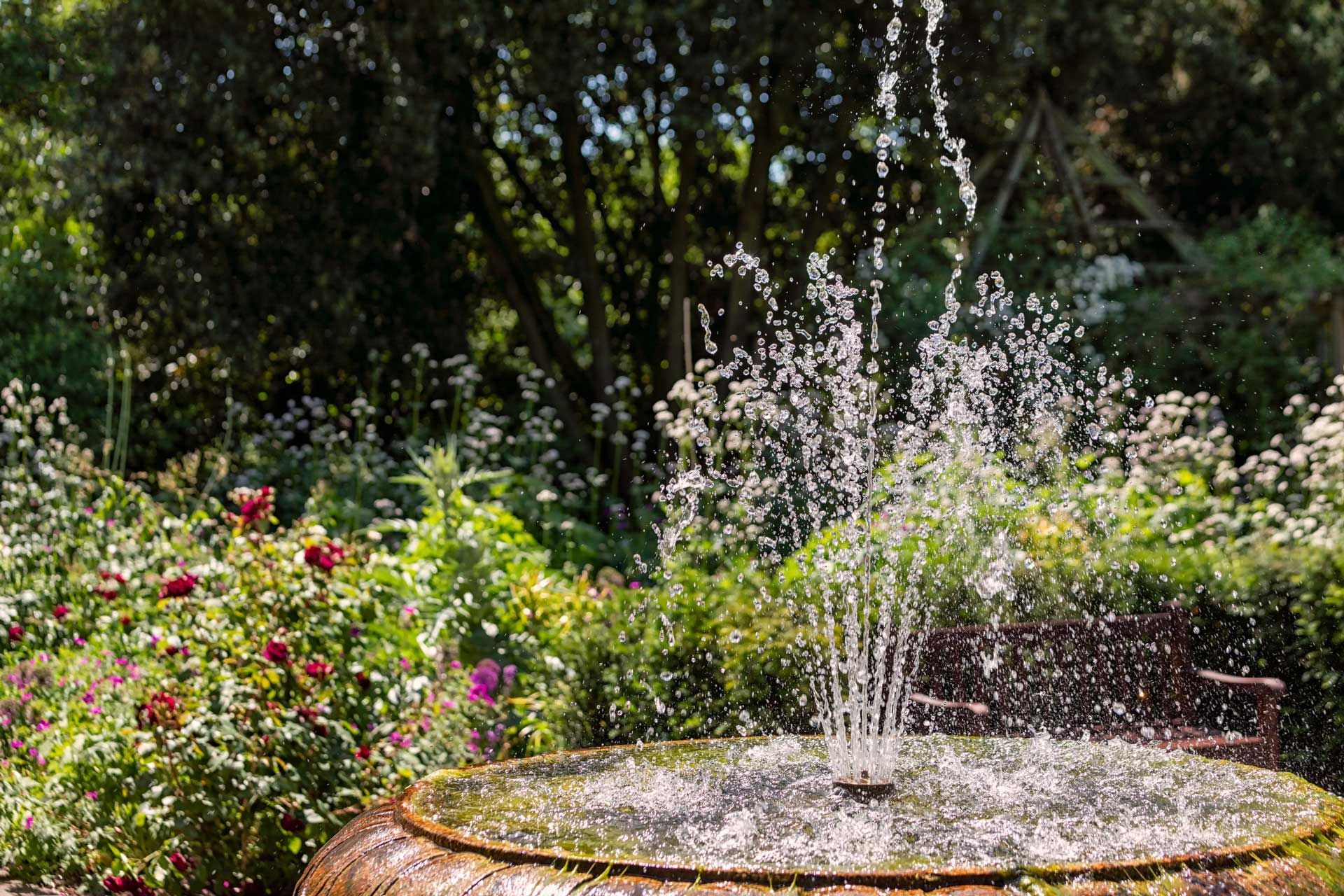 Fountain spraying water in a garden with flowers and trees.