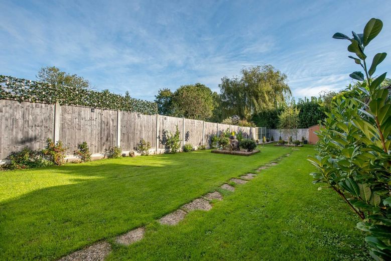 Green lawn in backyard, bordered by a wooden fence with plants and trees under a blue sky.