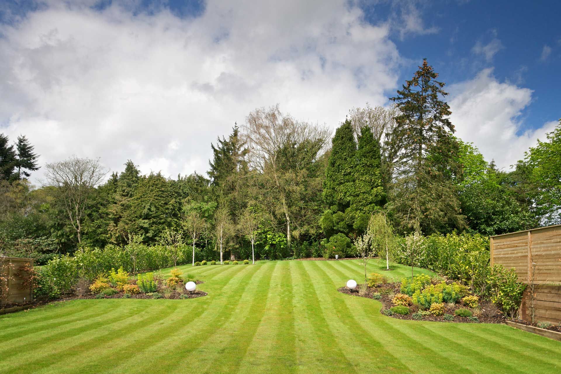 Lush green lawn with striped mowing pattern, bordered by flowerbeds, trees, and cloudy sky.