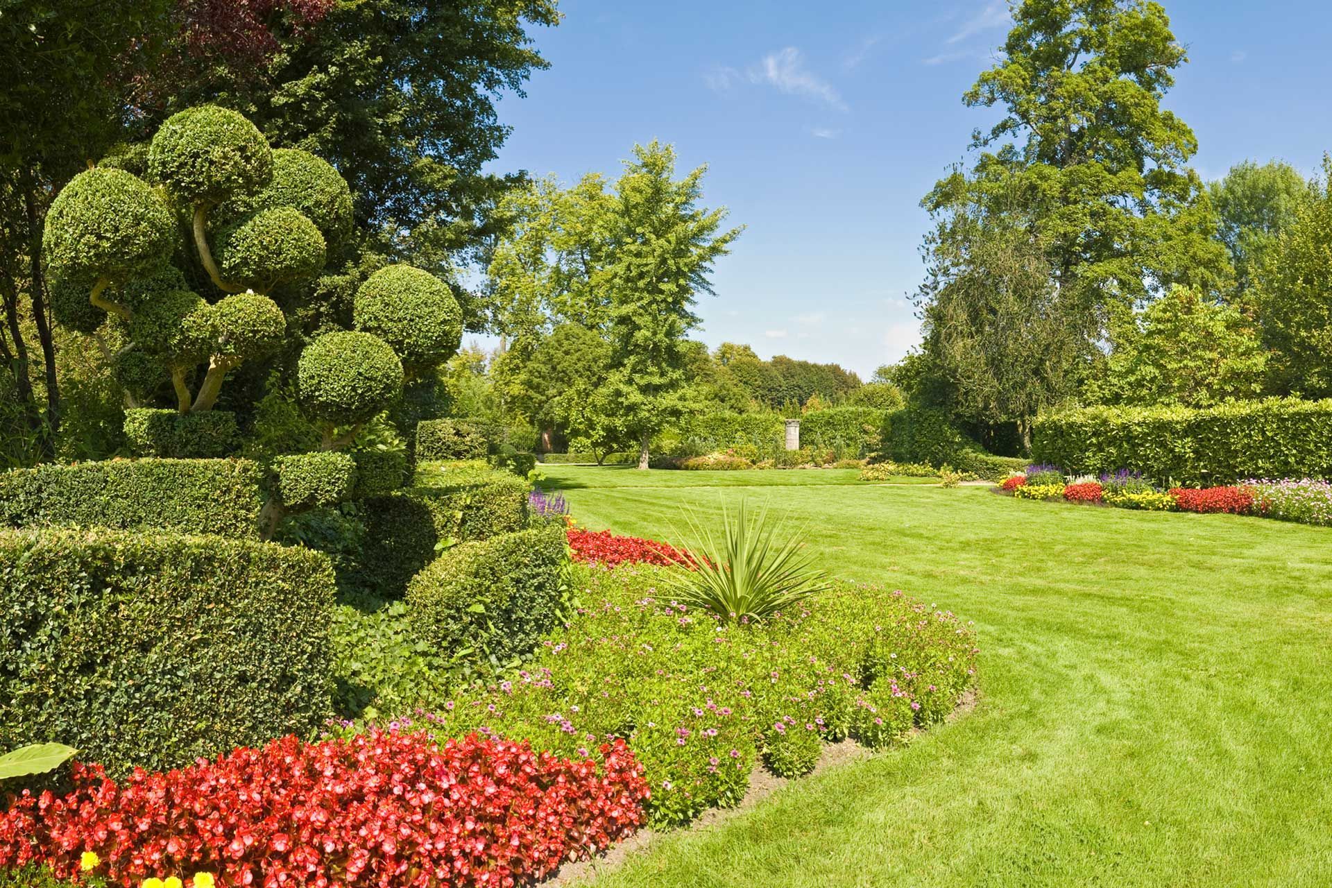 Manicured garden with green grass, hedges, and colorful flowers under a blue sky.