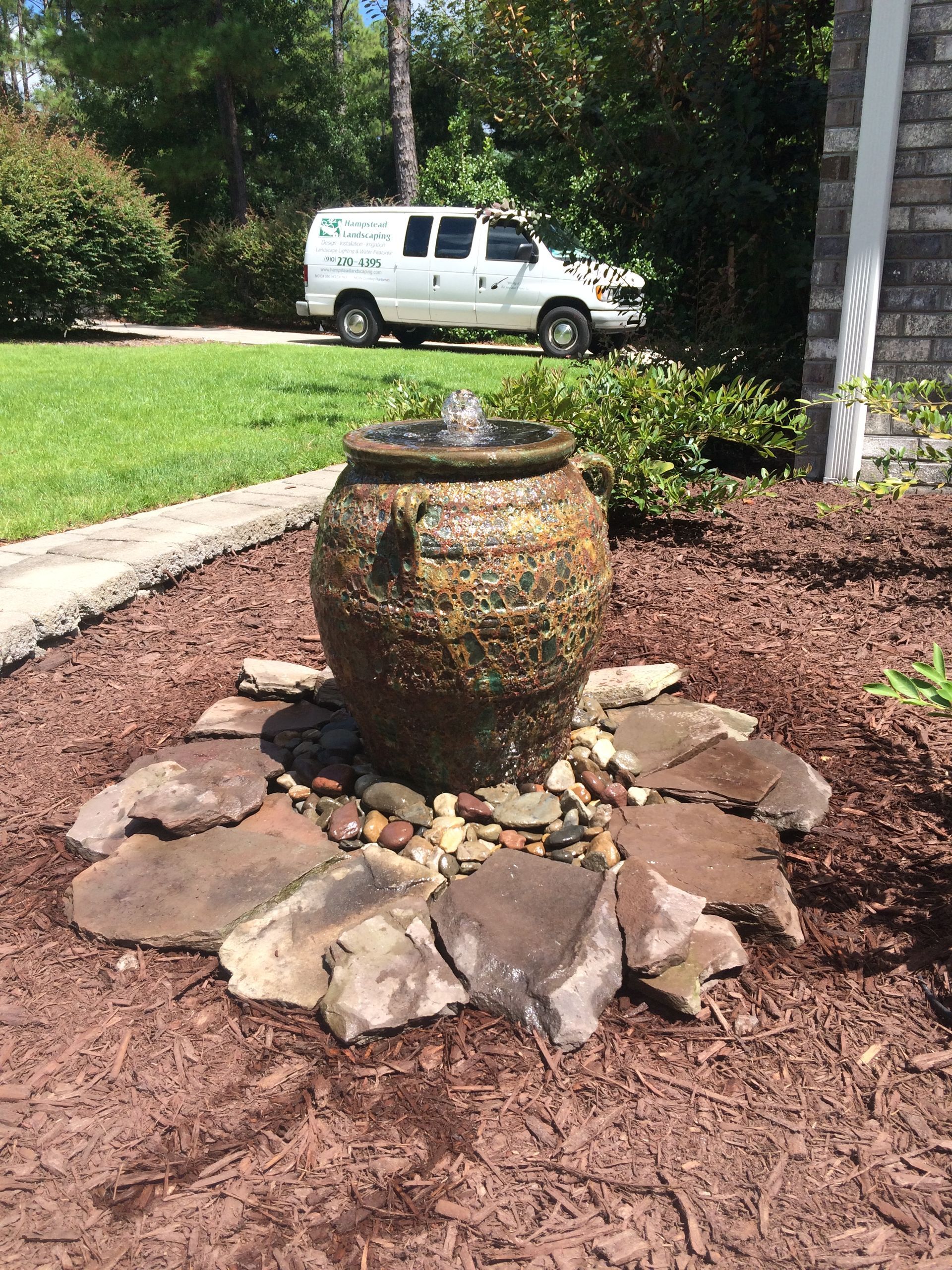 Stone fountain in a lush garden, with bright green plants and colorful flowers. Sunny day.