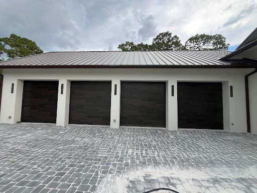 Modern garage with four dark doors, white exterior walls, and a gray paver driveway under cloudy skies