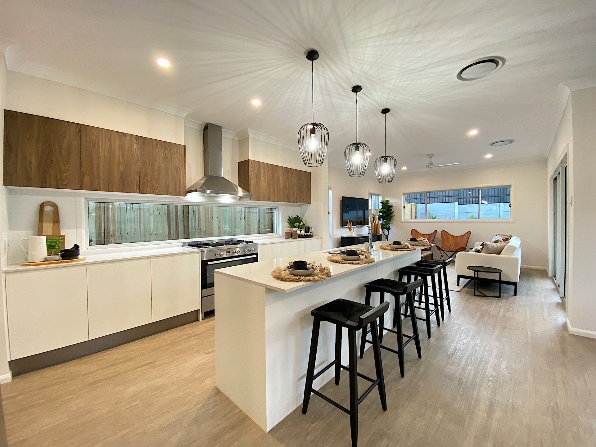 Modern kitchen with a white island, wood cabinets, and black bar stools; living area visible in the background.