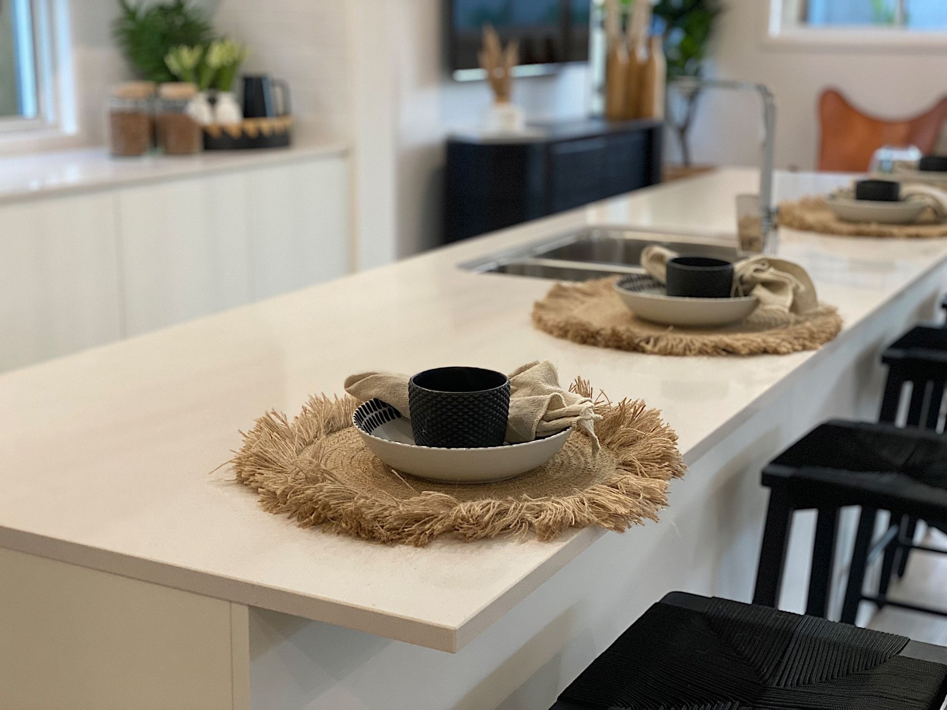 Kitchen island with two place settings: black mugs on beige placemats, white countertops, and black barstools.