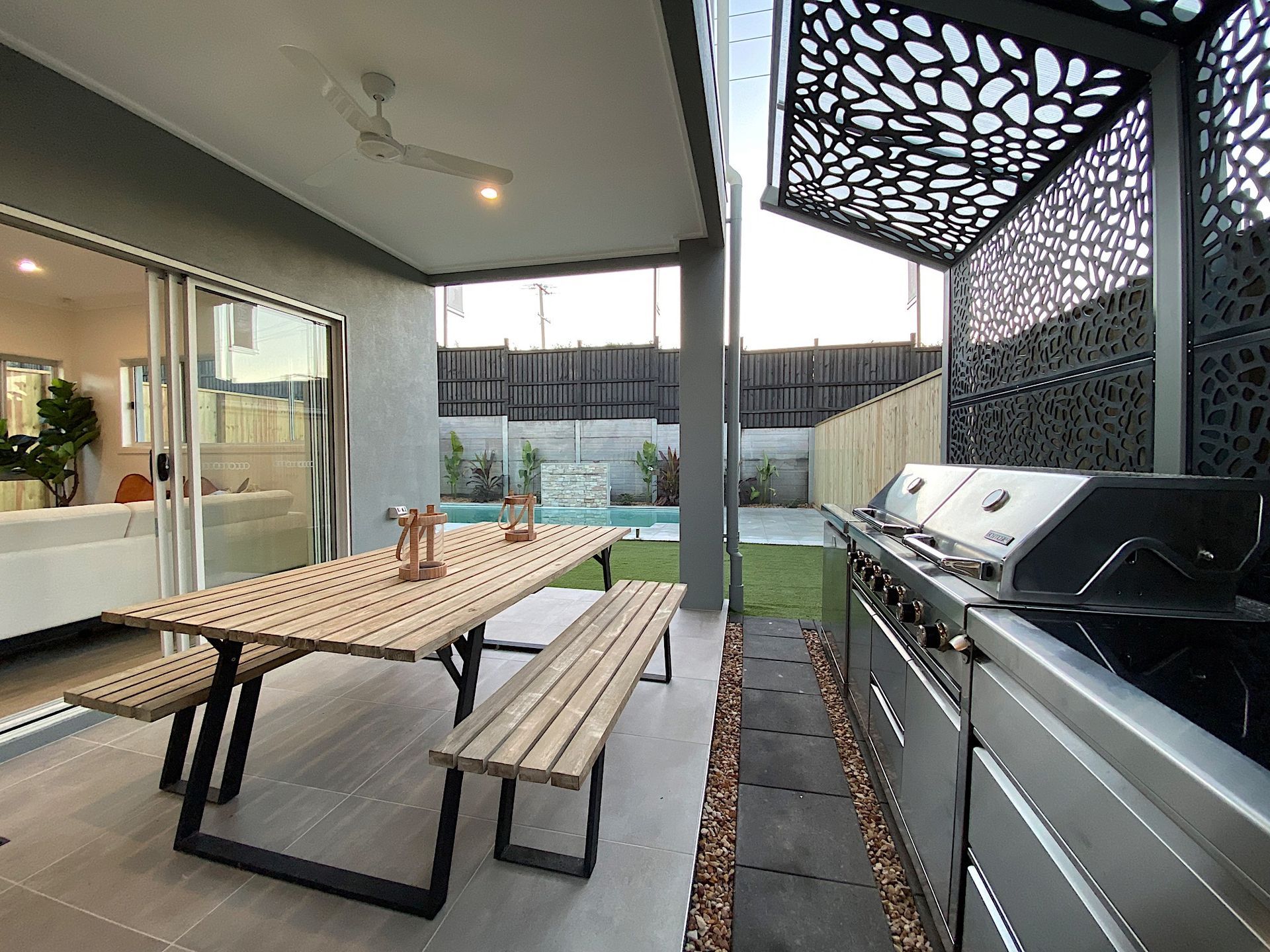 Outdoor patio with a wooden picnic table, stainless steel grill, and pool in the background.