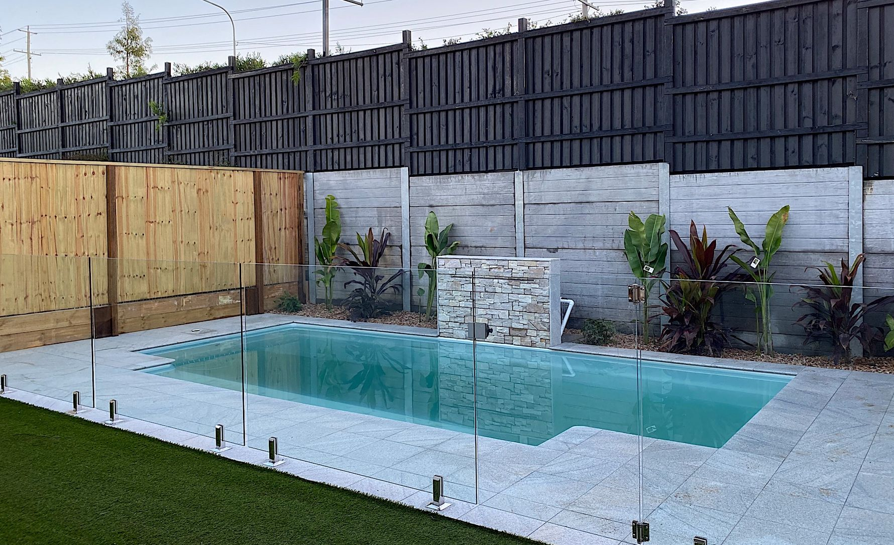 A rectangular pool surrounded by a stone patio and glass fencing, set against a wooden and dark gray fence.