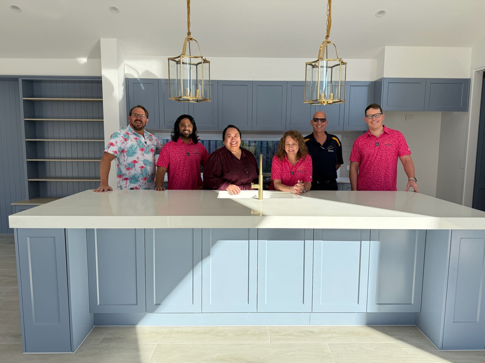 Six people pose in a light blue kitchen, smiling. They stand behind a large island, wearing pink shirts.