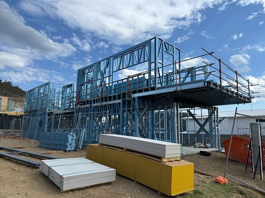 Steel frame of a house under construction, with scaffolding, building materials, and blue sky.